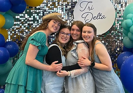 Eight smiling members of the Tri-Delta sorority pose with three large blue delta symbols on a green lawn.